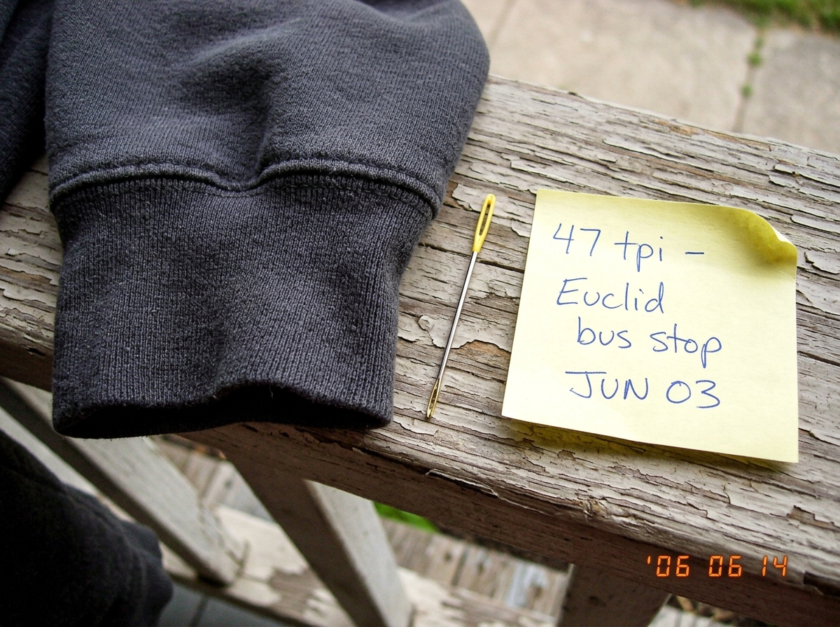A close-up photo of a gray hoodie cuff resting on a worn wooden porch railing, taken in natural light. A yellow sewing needle lies beside it for scale. A handwritten sticky note reads '47 tpi - Euclid bus stop JUN 03' in blue ballpoint pen. The weave is dense and visible, individual threads countabl