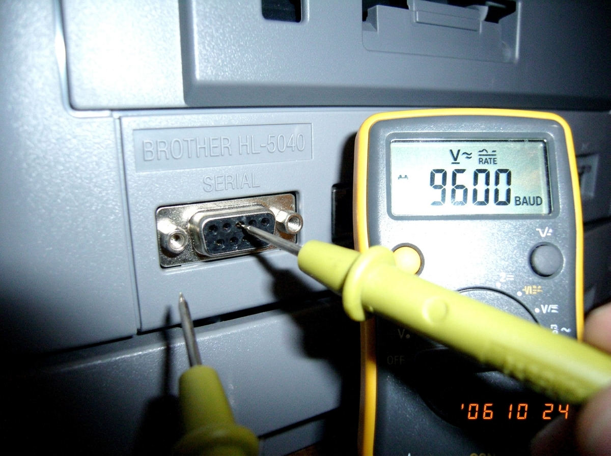 A close-up of a Brother HL-5040 label printer serial port with a multimeter probe touching pin 2, showing a reading of 9600 baud