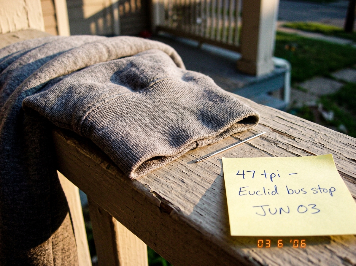 A close-up photograph of a worn gray hoodie cuff lying on a wooden porch railing in late afternoon sunlight. The weave is clearly visible - you can count 47 threads per inch by eye. A silver sewing needle rests next to the fabric for scale. A yellow sticky note in the corner has handwriting that say