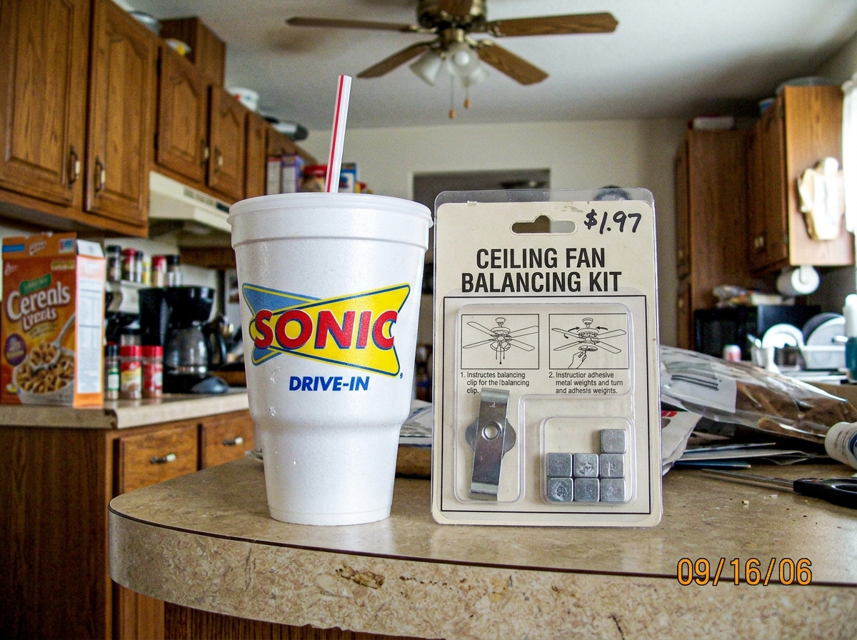 A white Sonic Drive-In cup sitting on a kitchen counter next to a ceiling fan balancing kit in its cardboard packaging. The cup has a straw and the Sonic logo visible. The balancing kit is a small clear plastic package labeled 'Ceiling Fan Balancing Kit' with a metal balancing clip and adhesive weig