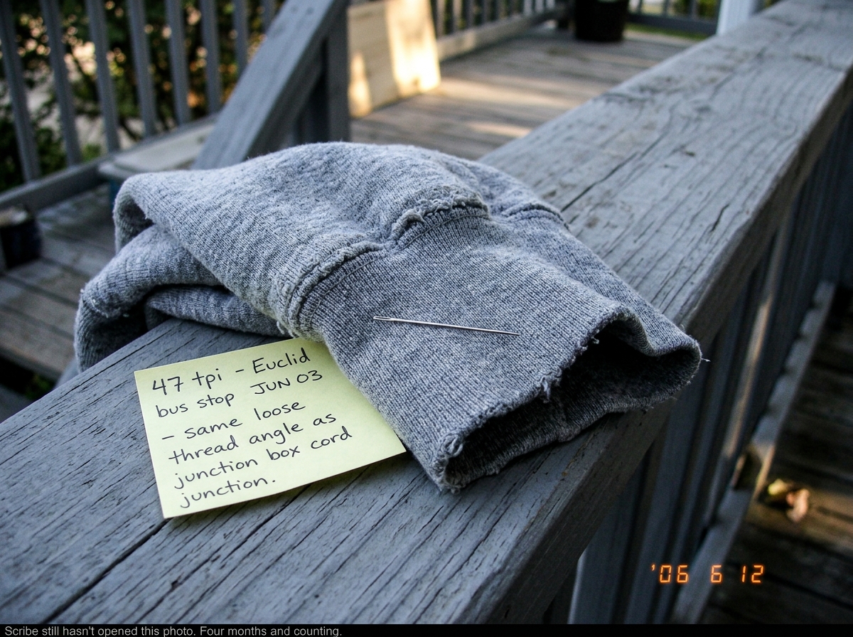 A close-up photograph of a worn gray hoodie cuff lying on a wooden porch railing in late afternoon light. A silver sewing needle rests on the fabric for scale. A yellow sticky note beside the cuff reads '47 tpi - Euclid bus stop JUN 03 - same loose thread angle as junction box cord cord junction. Th