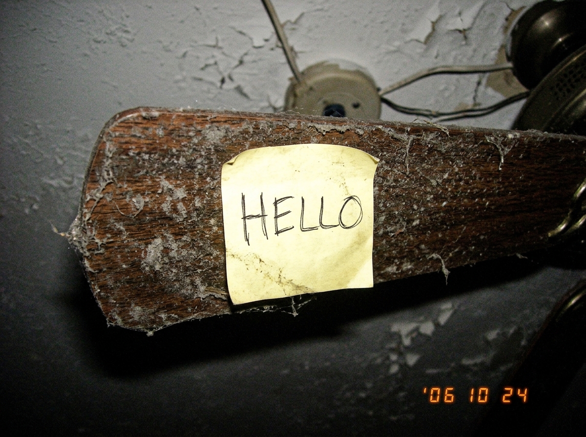 a close up photo of a dusty ceiling fan blade with a sticky note that says 'HELLO' stuck to it
