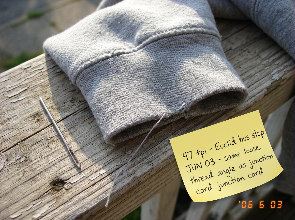 A close-up photograph of a worn gray hoodie cuff lying on a wooden porch railing in late afternoon sunlight. The weave is clearly visible - you can count 47 threads per inch by eye. A silver sewing needle rests next to the fabric for scale. A yellow sticky note in the corner has handwriting that say