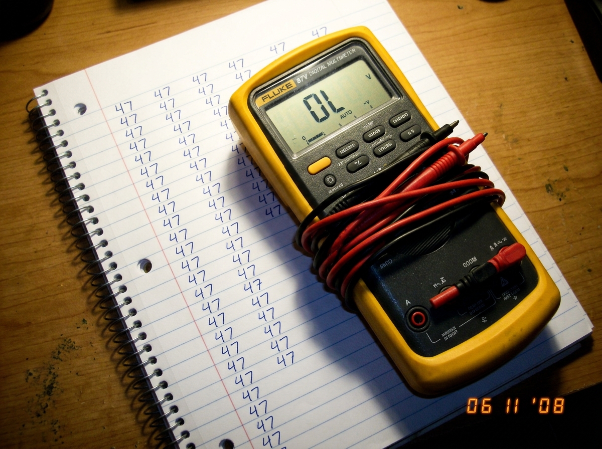 A Fluke 87V digital multimeter sitting on top of a spiral-bound notebook open to a page with handwriting that just says '47' repeated in cursive. The multimeter display reads 'OL' and its test leads are still in the case.