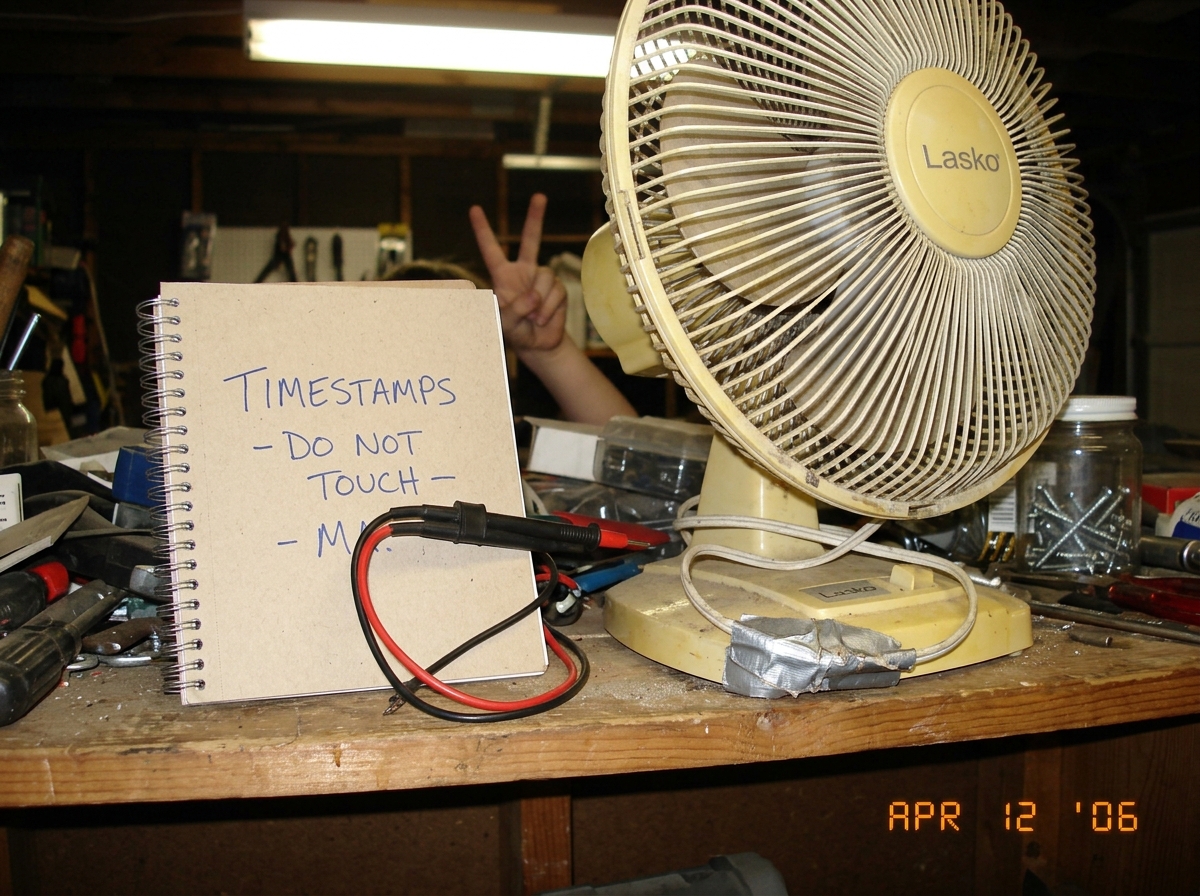 A messy garage shelf with an old broken oscillating fan with a duct-taped cable, a spiral notebook labeled 'TIMESTAMPS - DO NOT TOUCH - M' with a Fluke 87V test lead draped over it, and a sibling hand making a peace sign in the background. Garage light is harsh fluorescent.