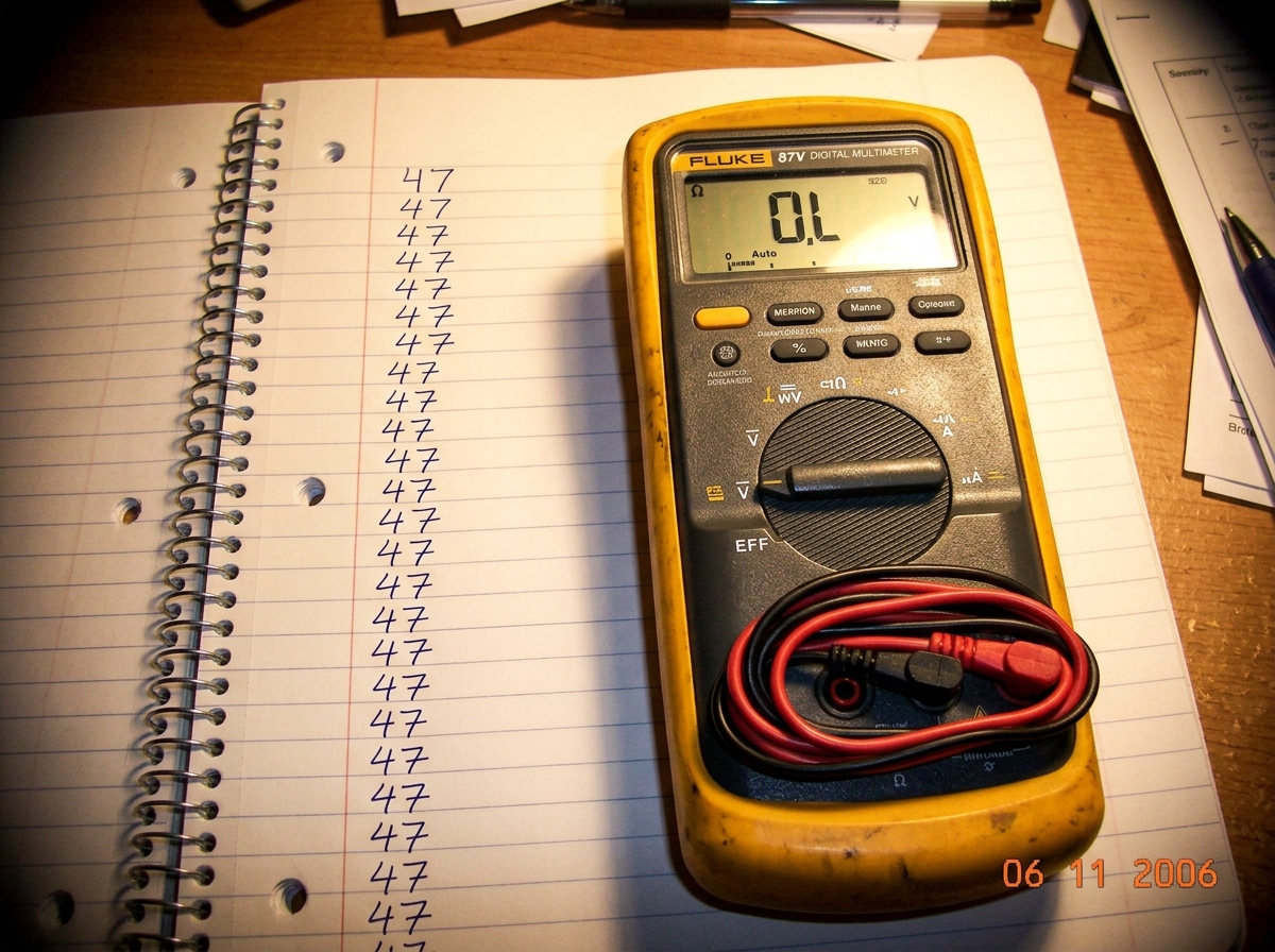 A Fluke 87V digital multimeter sitting on top of a spiral-bound notebook open to a page with handwriting that just says '47' repeated in cursive. The multimeter display reads 'OL' and its test leads are still in the case.