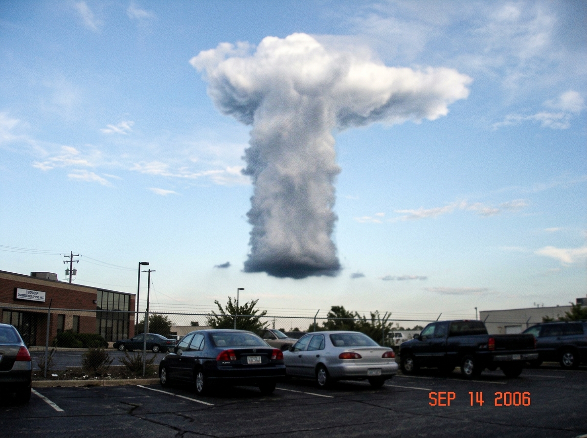 A grainy photo taken from an office parking lot looking up at a tall, column-like cumulonimbus cloud that rises vertically in a nearly straight stack, with flat base and distinctive anvil top, taken around late afternoon against a pale blue sky.