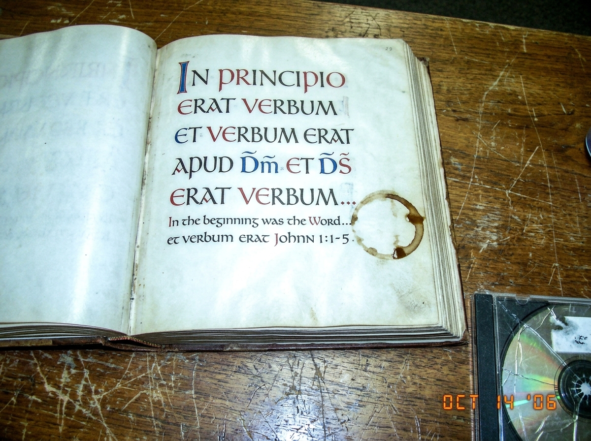 A blurry photo of a Lindisfarne Gospels facsimile page open to the Latin text of John 1:1-5, taken under harsh fluorescent light on a scratched wooden desk. A coffee mug ring stains the margin. A CD case is partially visible at the bottom edge, cracked.