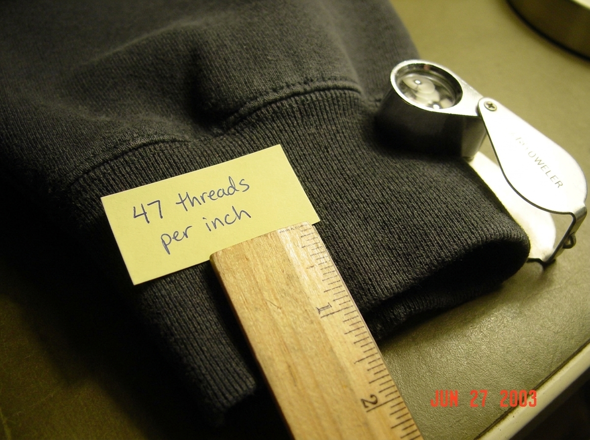 A close-up macro photograph of a dark gray hoodie cuff, taken with a small ruler laid across the fabric showing '47 threads per inch' handwritten in blue pen on a sticky note next to the edge. The weave is visible under magnification, with each thread clearly countable. In the corner, a small printe