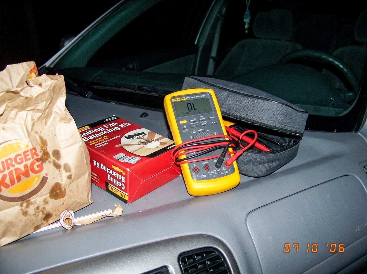 A Fluke 87V digital multimeter sitting on the dashboard of a parked car next to a ceiling fan balancing kit and a fast-food bag, the meter display reading 'OL' with test leads still coiled in the case