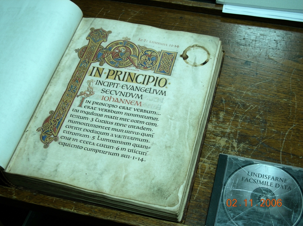 A blurry photo of a Lindisfarne Gospels facsimile page open to the Latin text of John 1:1-5, taken under harsh fluorescent light on a scratched wooden desk. A coffee mug ring stains the margin. A CD case is partially visible at the bottom edge, cracked.