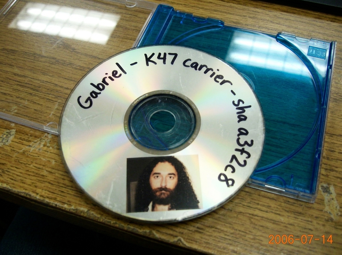 A close-up of a CD-R disc face with a handwritten label reading 'Gabriel - K47 carrier - sha a3f2c8' in black marker, the disc edge slightly scuffed, lying on a scratched desk next to a blue plastic CD jewel case half-open, fluorescent light reflecting off the disc surface