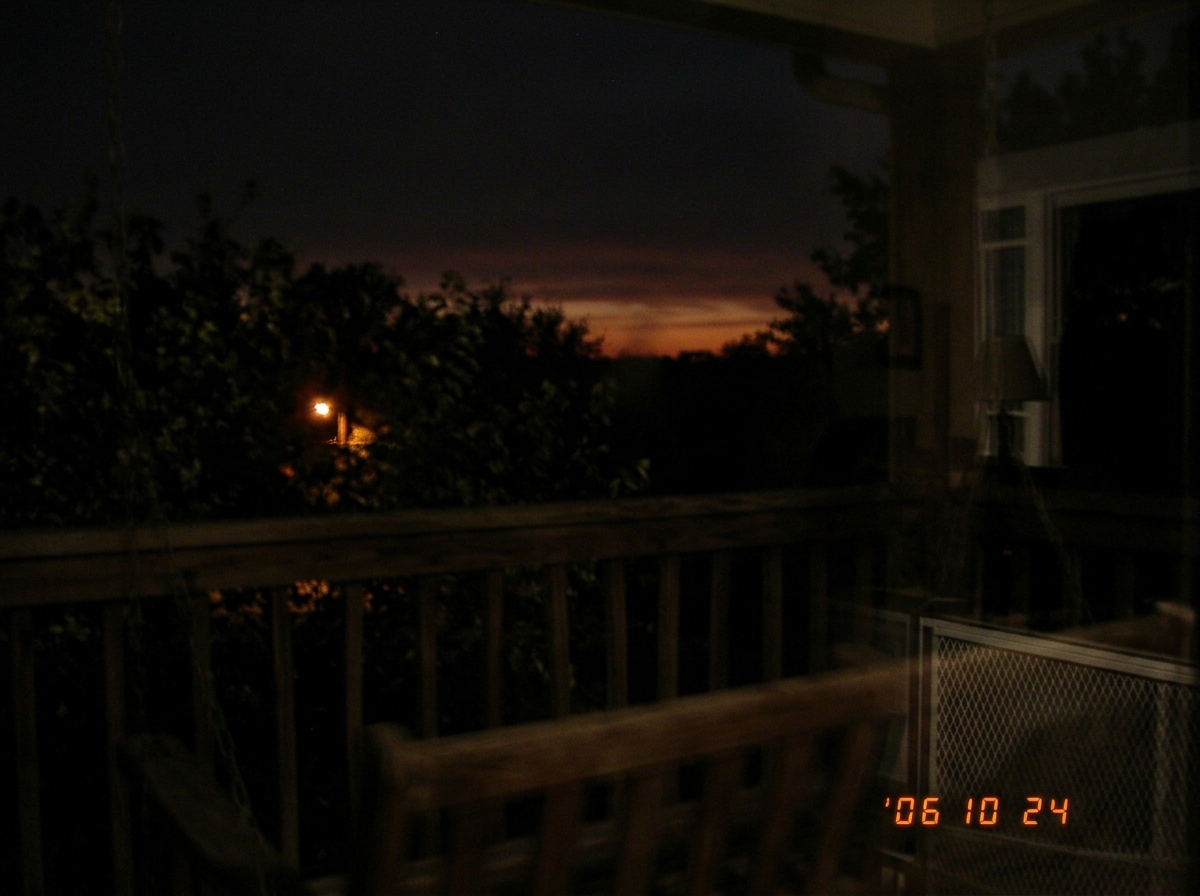 A blurry nighttime photo taken from a porch swing showing a dark horizon with an orange glow band fading. The view is partially obstructed by porch railing. A streetlight in the distance is barely visible through trees. The image is grainy, taken without flash, with screen door reflection visible in