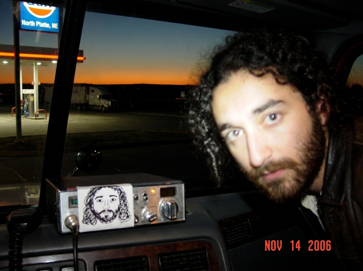 A blurry photo taken through a truck windshield at a rest area near North Platte, Nebraska at dusk. The sky has a thin orange band along the horizon. A gas station sign in the distance is partially visible. The dashboard shows a CB radio with a hand-drawn Gabriel sticker on it. Crooked frame, taken 