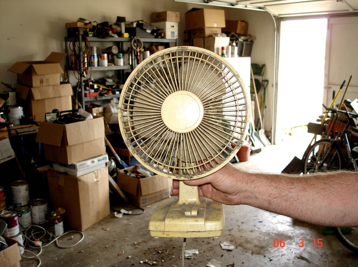 A dusty beige plastic fan with one blade missing, held aloft by a middle-aged man's hand in a cluttered garage, sunlight streaming through an open garage door onto boxes and shelving