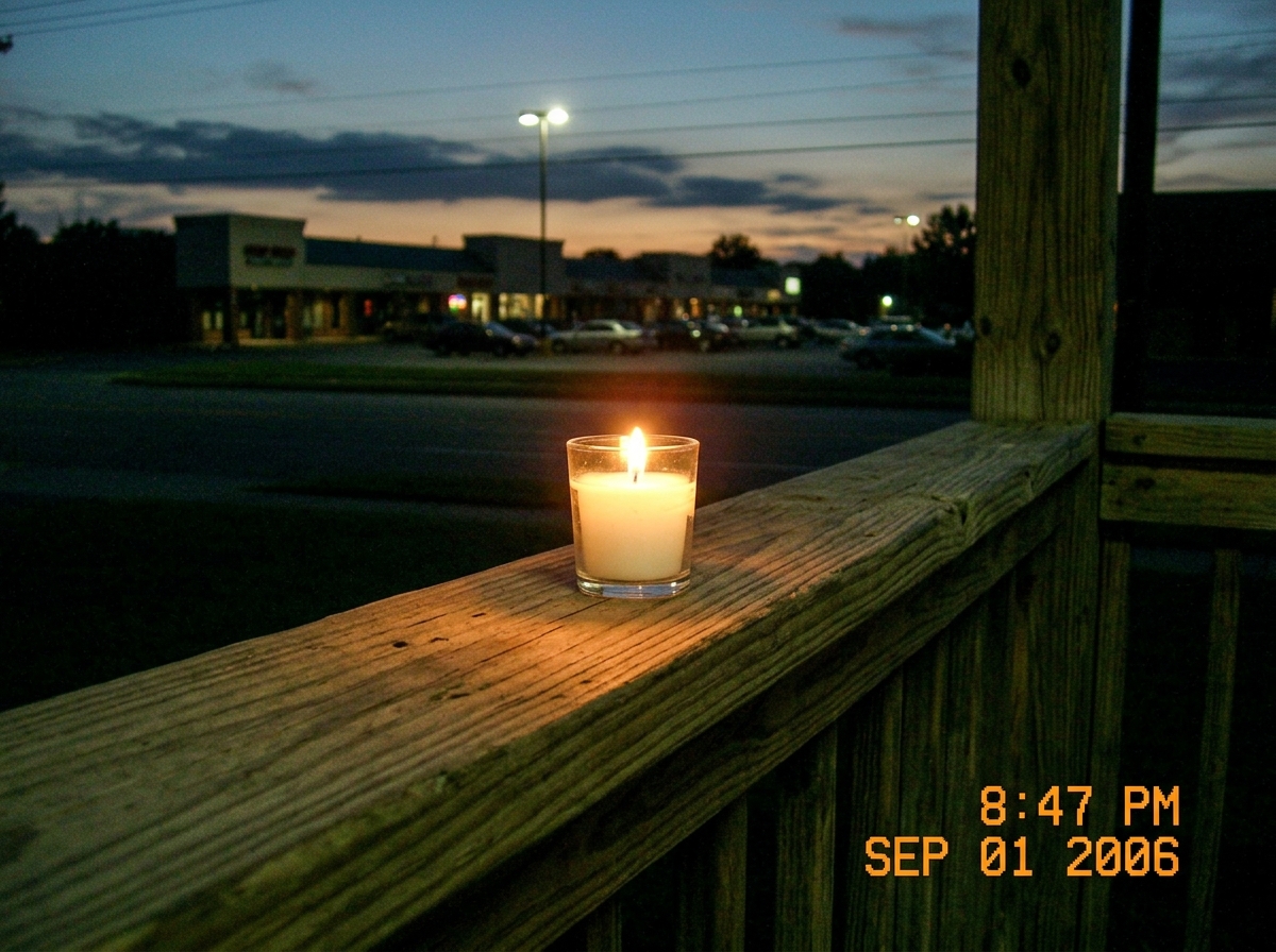 A close-up photo taken at night from a porch railing. A single white votive candle sits on a wooden rail, flame steady and upright casting a warm orange glow. In the background, a streetlamp and the silhouette of a strip mall are visible under a fading twilight sky. The camera timestamp info visible