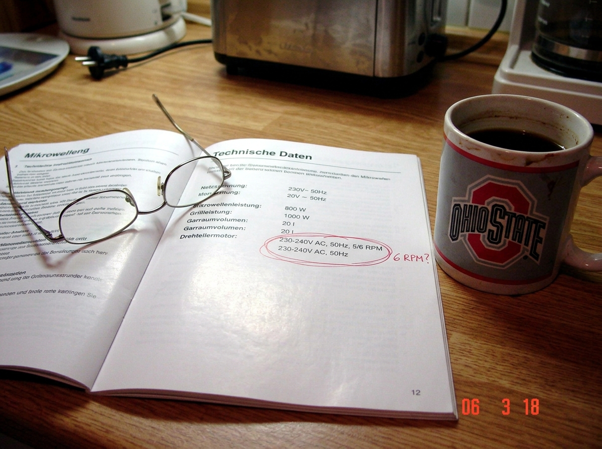 A German microwave instruction manual open to the technical specifications page. The turntable motor section is circled in red pen with '6 RPM?' written in the margin. The manual is in German, with words like 'Umdrehungen pro Minute' and 'Mikrowellenleistung' visible. A pair of reading glasses rests