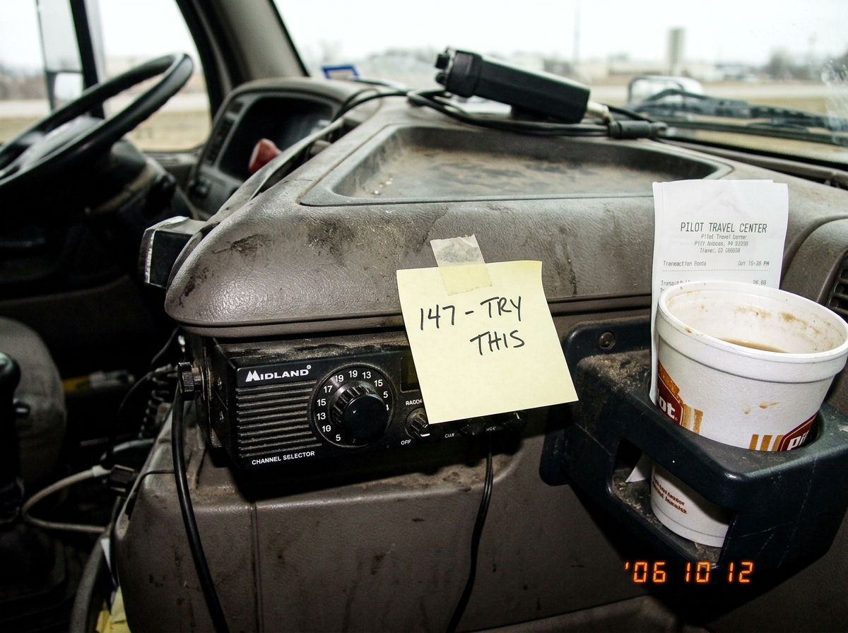 A close-up photo taken from a truck cab showing a CB radio mounted under the dashboard. The dial is tuned to channel 19, but a sticky note is taped next to it reading '147 - TRY THIS' in black pen. The dashboard is dusty, with a half-empty coffee cup in the cup holder and a crumpled receipt from a P