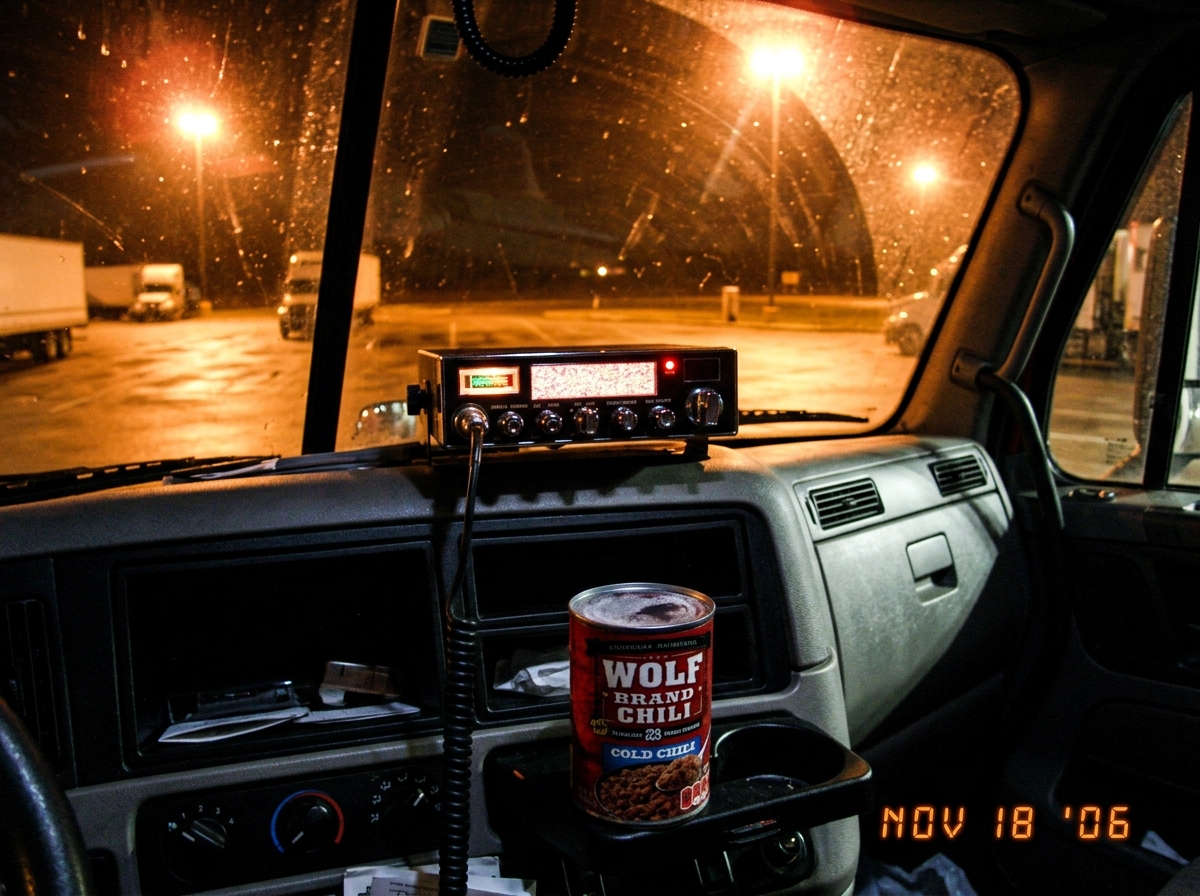 A dashboard-mounted CB radio at night with static on the display, a can of Wolf Brand chili sitting cold on the center console, sodium light casting orange glow through the windshield of a parked semi truck