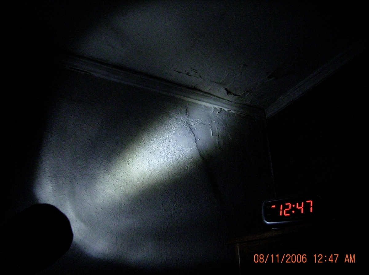 A dark bedroom ceiling seen from below, flashlight beam cutting across plaster, late night timestamp 12:47 AM visible on a digital clock in the corner