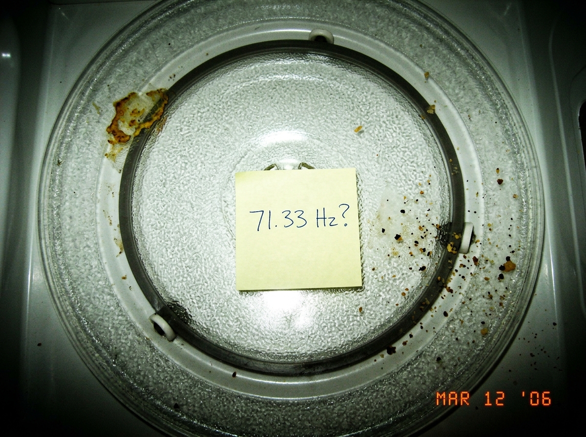 A microwave turntable seen from above with a small handwritten sticky note stuck to the glass plate reading '71.33 Hz?' in blue ink. A few crumbs and a dried mustard stain are visible on the turntable rim. The photo is taken under harsh kitchen fluorescent light.