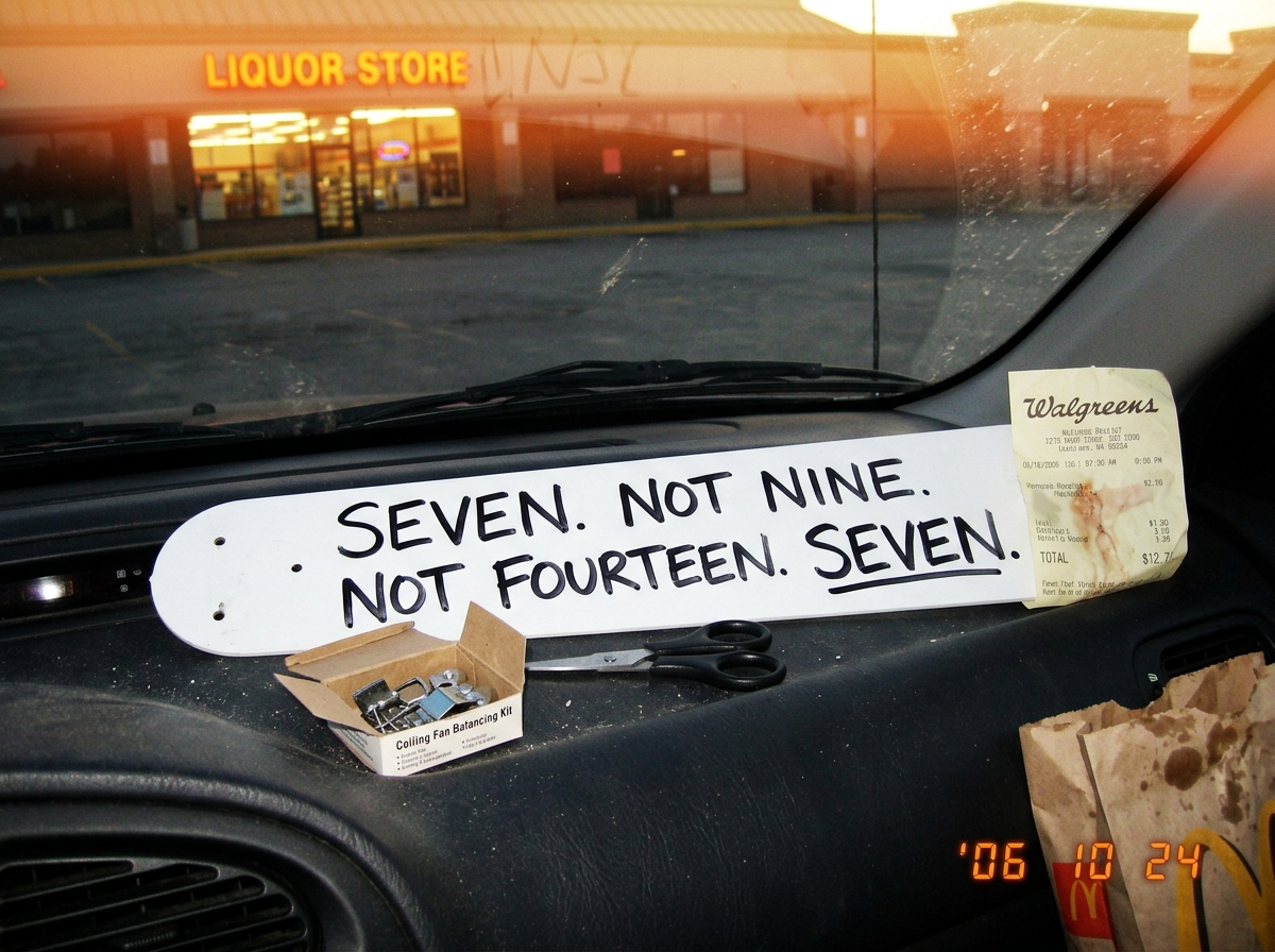 A close-up photograph of a single white ceiling fan blade lying on a car dashboard. Written on it in black sharpie, in large block letters: 'SEVEN. NOT NINE. NOT FOURTEEN. SEVEN.' Next to the blade sits a crumpled Walgreens receipt with the total partially visible. A pair of scissors and a small bal