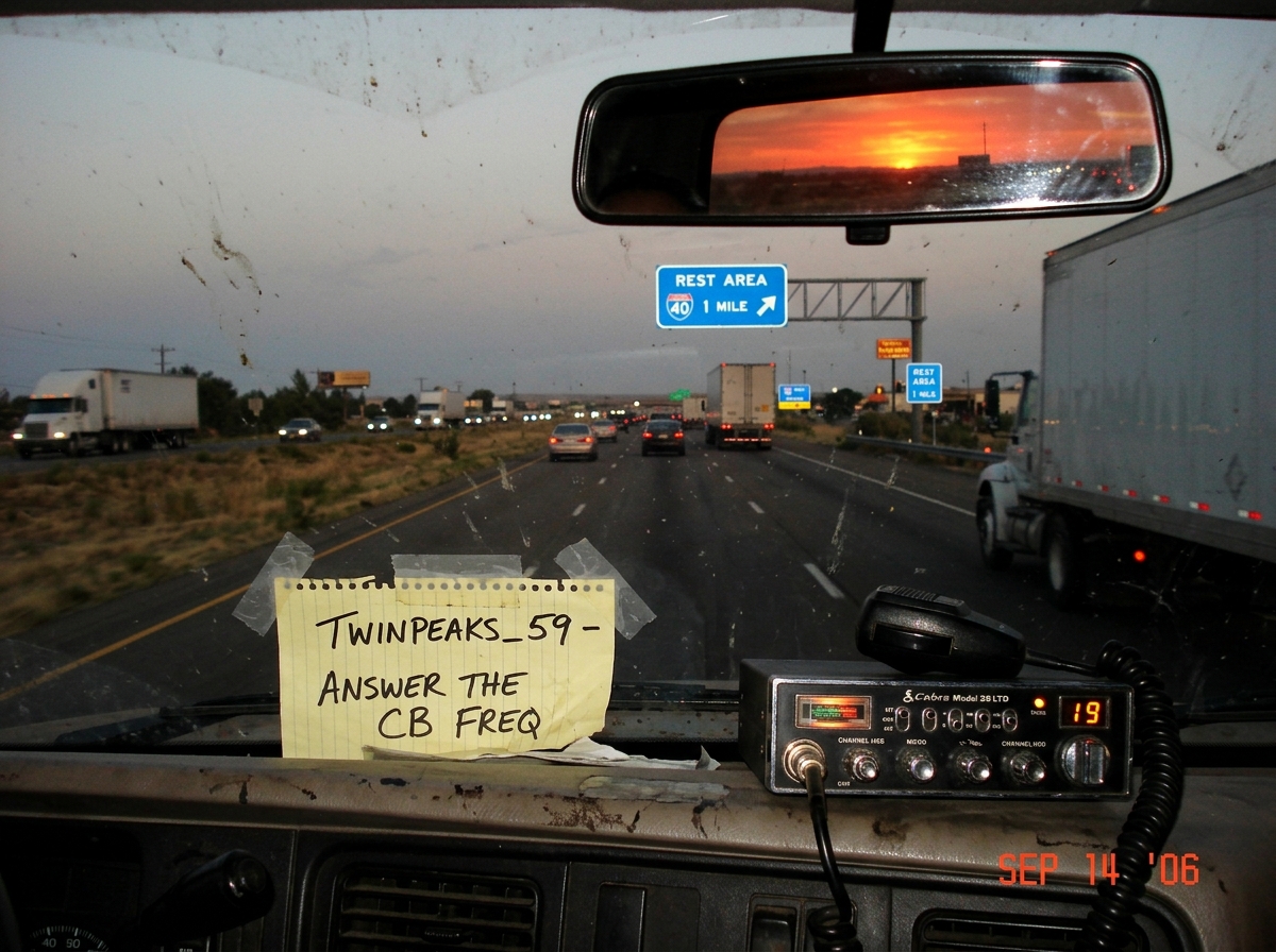 A photo taken through a dusty truck windshield showing a highway sign for a rest area ahead. On the dashboard, a piece of paper with 'TWINPEAKS_59 - ANSWER THE CB FREQ' written in black marker is taped next to a CB radio. The sun is setting orange in the rearview mirror reflection.