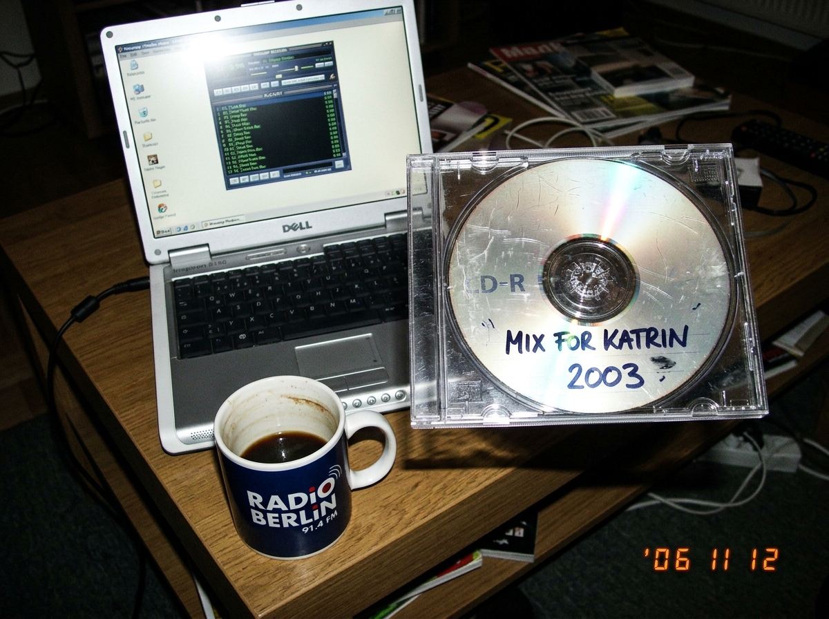 photograph of a scratched CD-R on a coffee table next to a laptop showing a Winamp playlist with FLAC files, the CD has a handwritten label 'MIX FOR KATRIN 2003', the coffee mug has a Berlin radio station logo