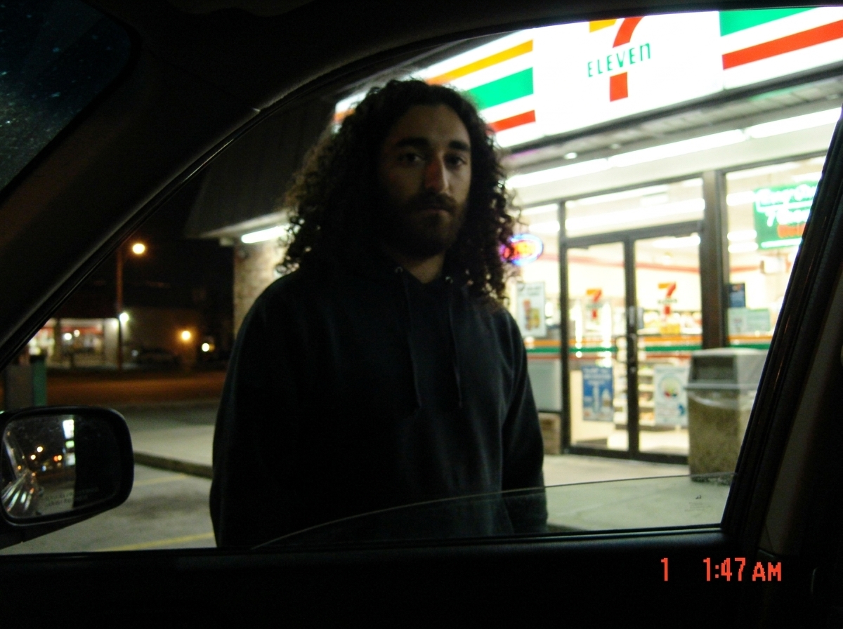 A blurry photo of a 7-Eleven storefront at night, taken from a car window. The timestamp in the corner reads '1:47 AM'. A single figure in a hoodie stands near the entrance, face obscured by shadow and the store's neon glow.