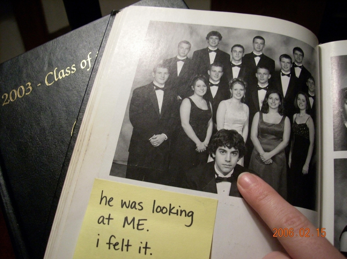 A scanned yearbook page from 2003 showing a high school group photo. A dark-haired boy in the corner is slightly turned toward the camera while the rest of the group faces forward. A woman's finger points at him from the edge of the scan. A handwritten sticky note below reads 'he was looking at ME. 