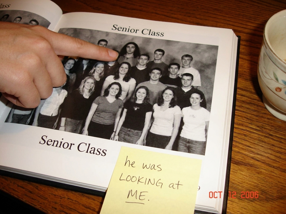 Scanned 2003 yearbook page open to the senior class group photo. A young woman's finger points at a dark-haired boy in the back corner who appears to be looking slightly off-camera. A handwritten sticky note below reads 'he was LOOKING at ME.' A teacup is visible at the edge of the table.