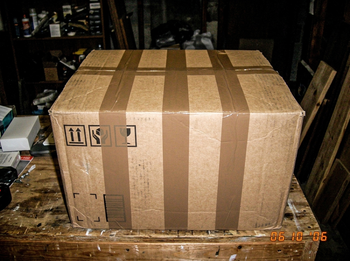 A close-up photo of a cardboard shipping box with packing tape applied in perfect, symmetrical parallel lines, taken with a cheap digital camera flash