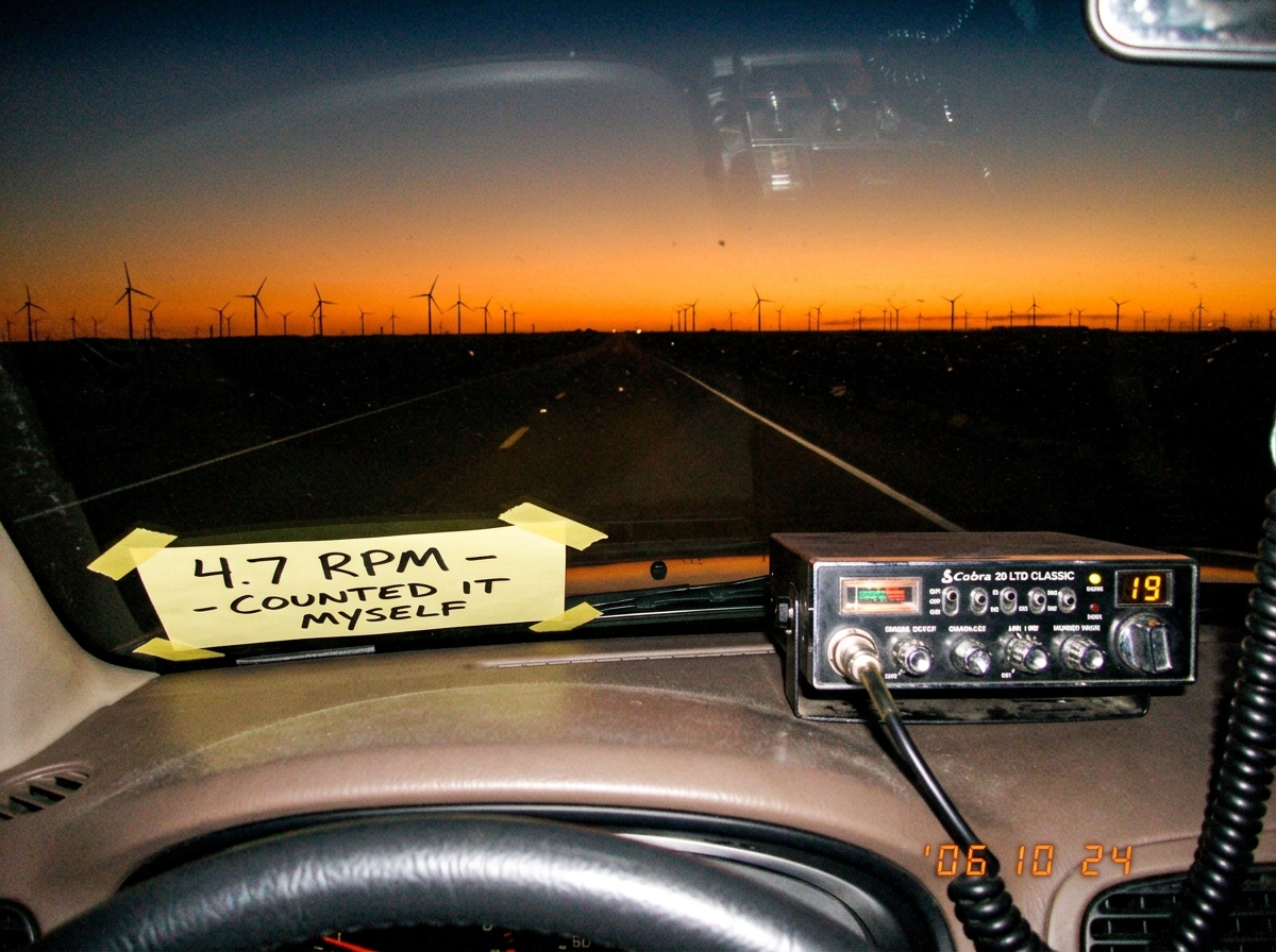 A windshield view of a long straight highway at dusk with dozens of wind turbines on the horizon, their blades silhouetted against an orange sunset sky. A CB radio sits on the dashboard, partially in frame. A handwritten sticky note on the windshield says '4.7 RPM - COUNTED IT MYSELF' in thick black