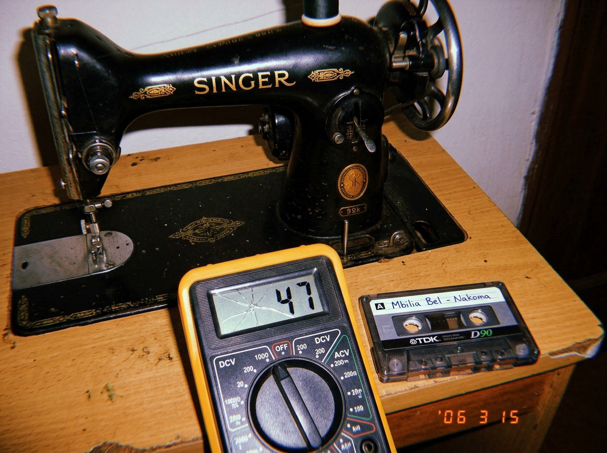 A cheap digital multimeter with a cracked LCD screen showing '47' next to a Singer sewing machine and a cassette tape labeled 'Mbilia Bel - Nakoma'
