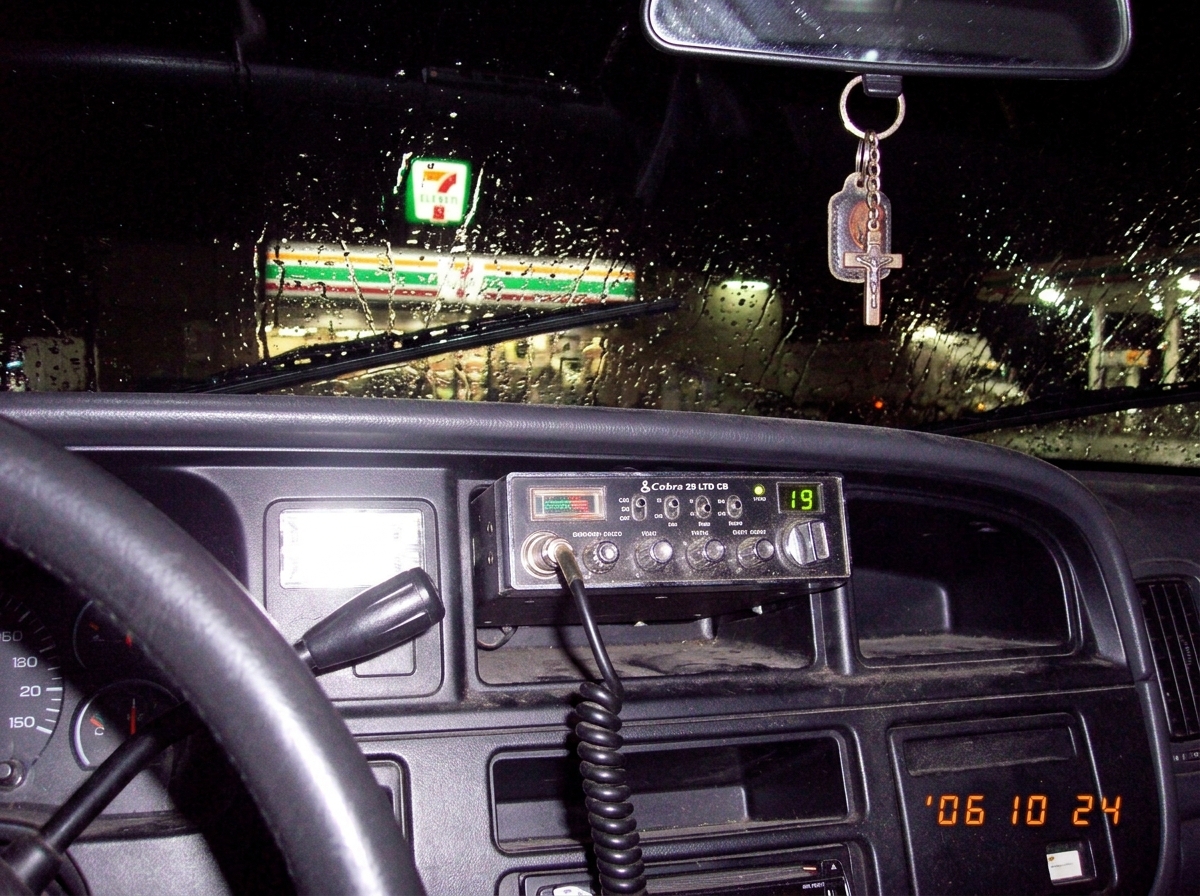 A close-up photo taken from the inside of a truck cab, showing a CB radio mounted under the dashboard. The display shows 'CH 19' with a faint green glow. Beyond the windshield, a 7-Eleven store sign glows in the distance at night, partially obscured by rain streaks on the glass. A keychain with a sm