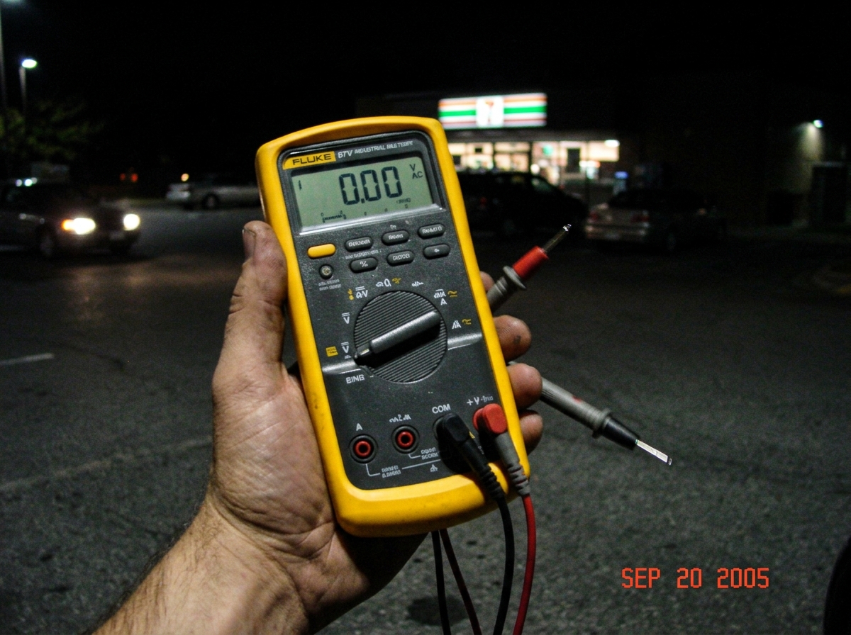 A blurry nighttime photo taken with a cheap digital camera of a Fluke 87V multimeter held in someone's hand, the display shows 0.00V with the leads plugged in but not touching anything. Behind the meter, a dimly lit parking lot has a 7-Eleven store sign visible in the far background, date stamp show