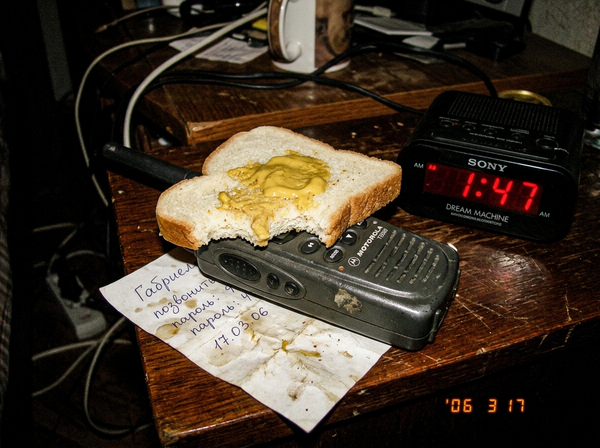 A close-up photo of a half-eaten slice of white bread with a glob of yellow mustard on it, sitting on top of a small handheld walkie talkie radio. A digital alarm clock beside it reads '1:47 AM' in red LED digits. A crumpled piece of paper with Cyrillic handwriting lies underneath, partially visible