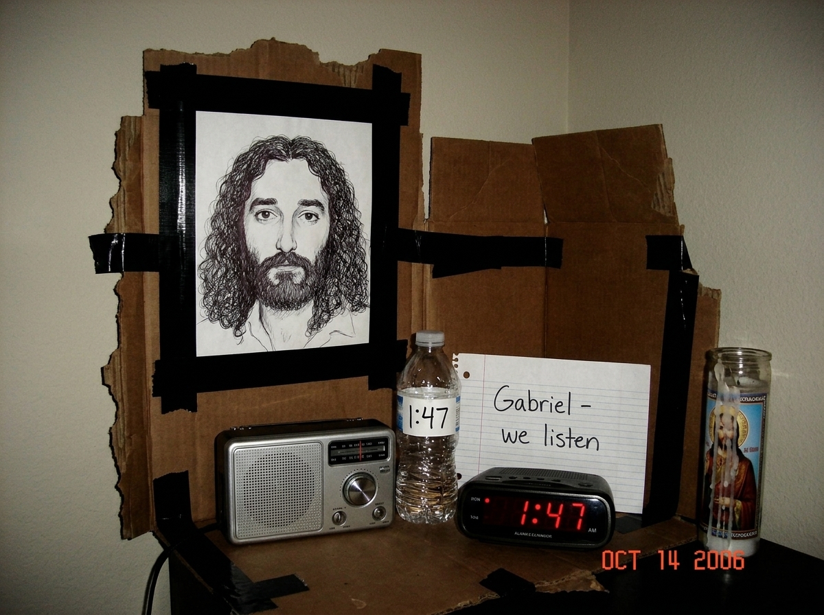 A crude shrine made from cardboard and electrical tape, with a hand-drawn picture of Gabriel taped to a wall, a small analog radio tuned to static, a plastic water bottle labeled '1:47', and a digital alarm clock showing 1:47 AM with red LED digits. A handwritten note reads 'Gabriel - we listen'. A