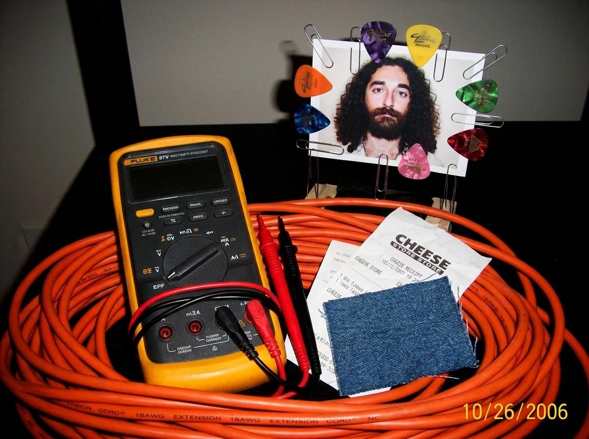 A photo of a Fluke 87V multimeter sitting on a coil of 16AWG extension cord next to a cheese receipt and a denim fabric swatch, with a small Gabriel Chantayan shrine made of paperclips and guitar picks in the background