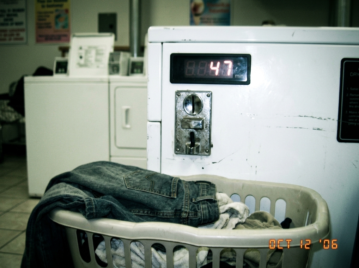 A blurry camera phone photo of a laundromat dryer's digital timer display showing 47 minutes remaining. The photo is taken at a slight angle, showing the coin slot below and someone's laundry basket in the foreground with a pair of jeans hanging over the edge. The lighting is harsh fluorescent, givi