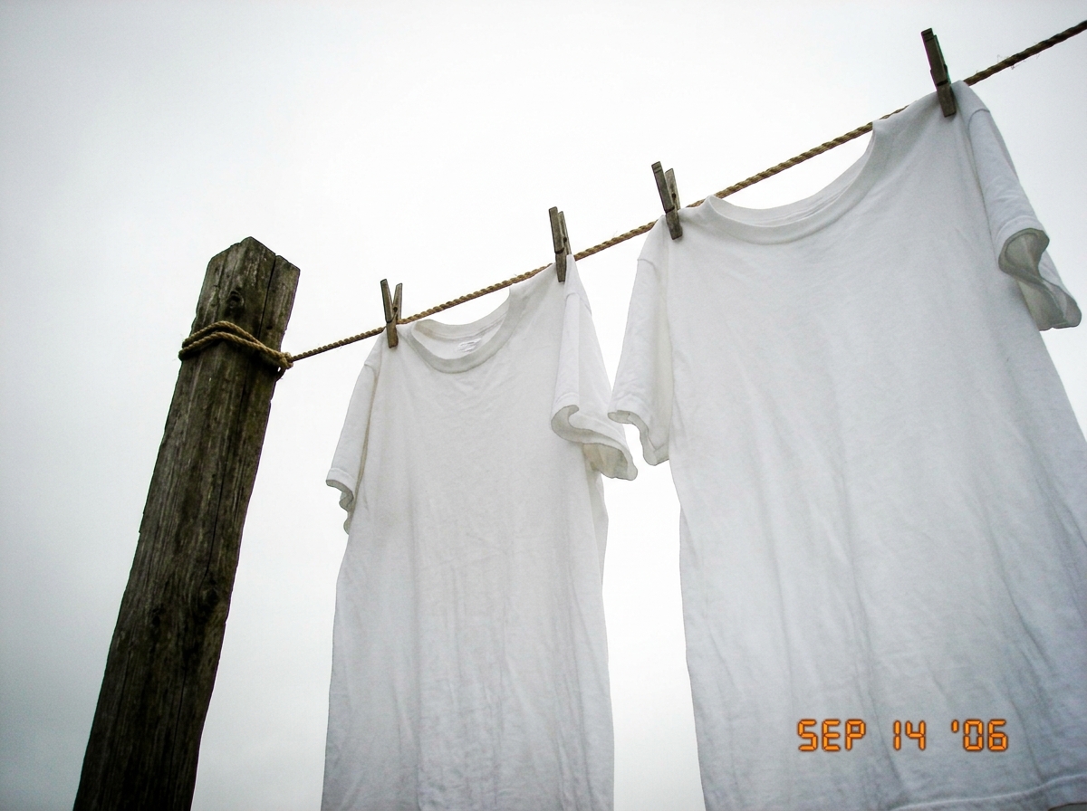 A photograph of an empty wooden clothesline against a grey overcast sky, with two white shirts hanging limp and motionless. The image is slightly overexposed, making the sky look almost white, blending with the shirts so it's hard to tell where fabric ends and atmosphere begins. Shot from below look