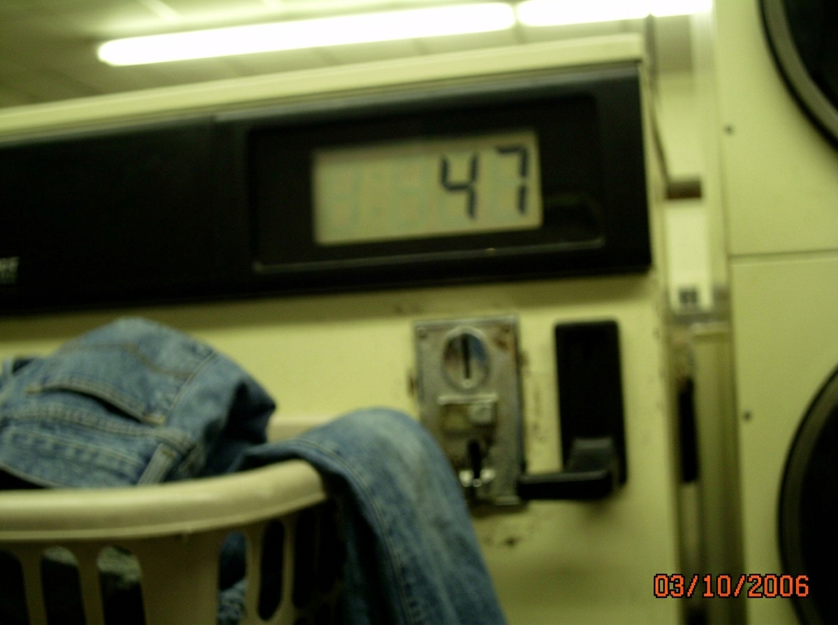 A blurry camera phone photo of a laundromat dryer's digital timer display showing 47 minutes remaining. The photo is taken at a slight angle, showing the coin slot below and someone's laundry basket in the foreground with a pair of jeans hanging over the edge. The lighting is harsh fluorescent, givi