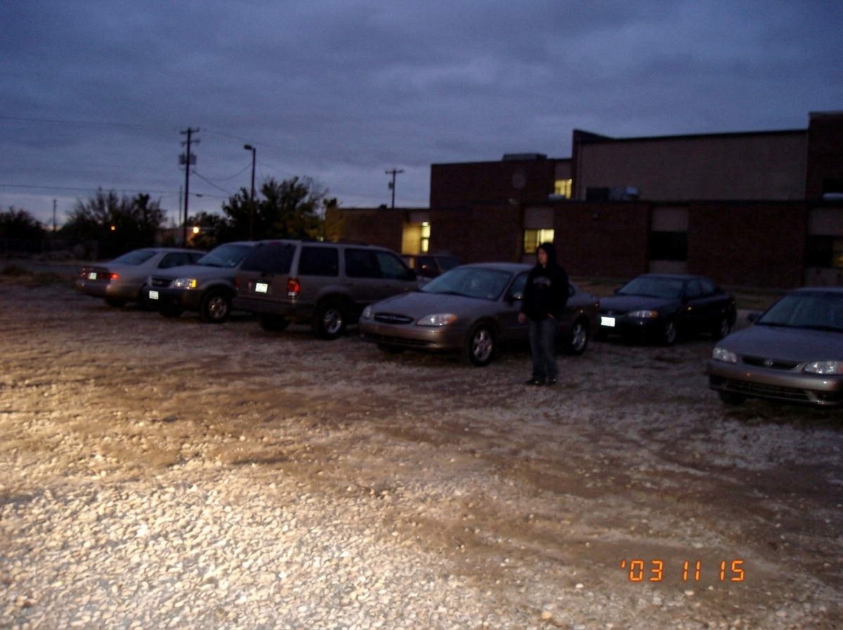 A blurry, dusk-lit photo of a gravel parking lot behind a high school, taken from across the lot. A lone figure in a hoodie stands near a row of parked cars, half-obscured by shadows. The image is grainy and slightly tilted, dated 2003 watermark in the corner.