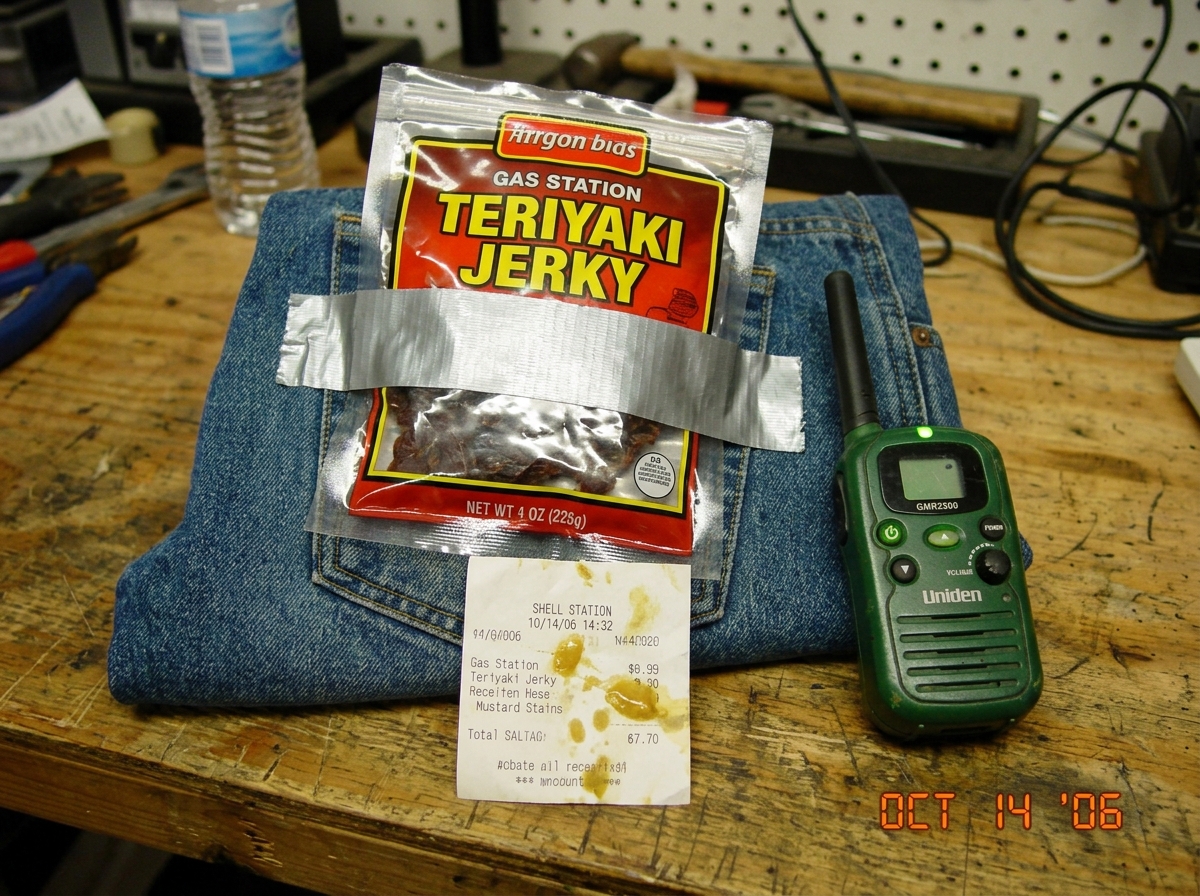 Shrine photo of a gas station beef jerky bag taped to a piece of denim with a small green LED walkie talkie next to it, mustard-stained receipt visible