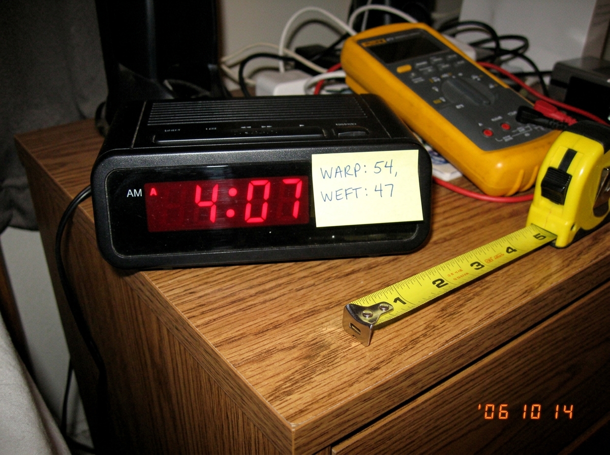 A photo of a digital alarm clock on a nightstand showing 4:07 AM. Next to it lies a yellow tape measure partially unspooled, its metal tip resting at the 1.47 inch mark. A sticky note on the clock reads 'WARP: 54, WEFT: 47' in neat handwriting. A Fluke 87V multimeter is visible in the background.
