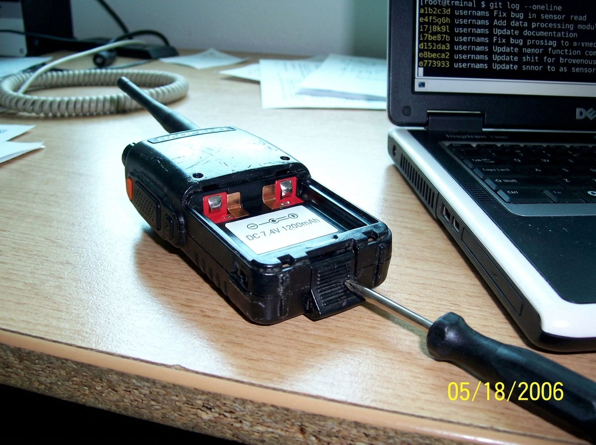 A photo of the same model Baofeng UV-5R walkie-talkie on a desk, with the battery compartment OPEN showing the battery contacts and a label reading 'DC 7.4V 1200mAh', and next to it a Dell laptop running a terminal with 'git log --oneline' output visible. A screwdriver rests on the battery release l