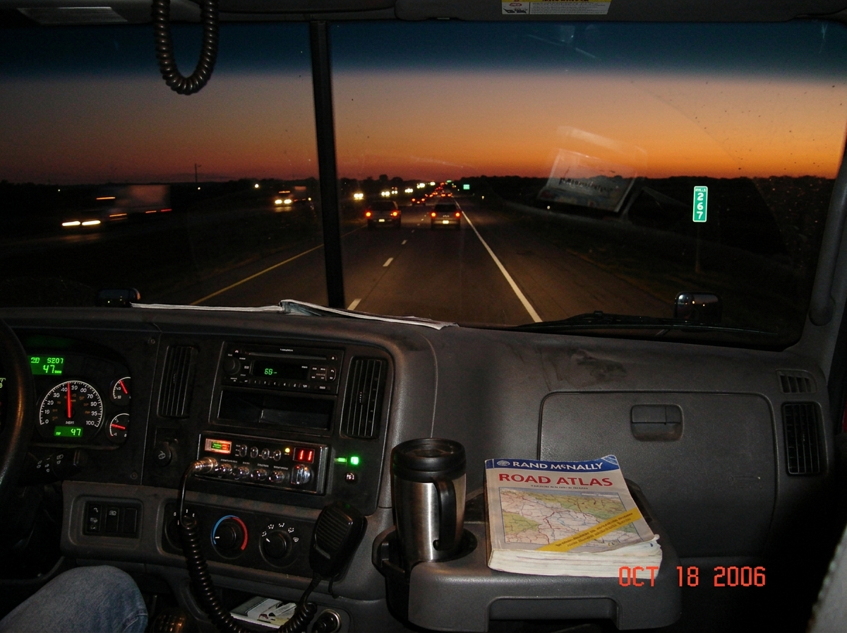 A photo taken from inside a truck cabin at dusk, looking out through the windshield at mile marker 267 on a long straight highway. The sky is a deep orange and purple fading to dark blue. The dashboard has a CB radio mounted below the radio, its red channel display glowing '11' and the green 'TX' li