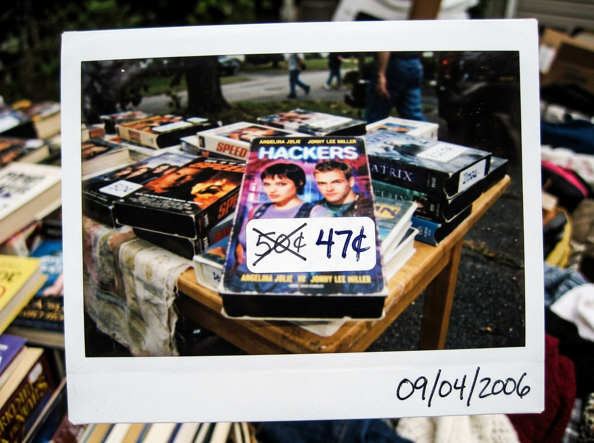 A blurry Polaroid of a yard sale table with a stack of VHS tapes. The price sticker on a copy of 'Hackers' is clearly visible but the number '50' is crossed out and replaced with '47' in messy handwriting. The date '09/04/2006' is scribbled in the corner.