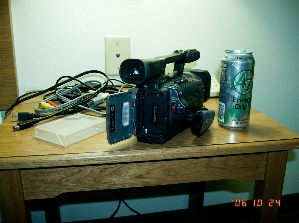 A Sony DCR-VX2000 camcorder with its battery compartment open, sitting on a table cluttered with cables and a half-empty can of 47-brand energy drink. The camcorder's serial number sticker is clearly visible but intentionally blurred out. The background shows a generic motel room with a Euclid socke