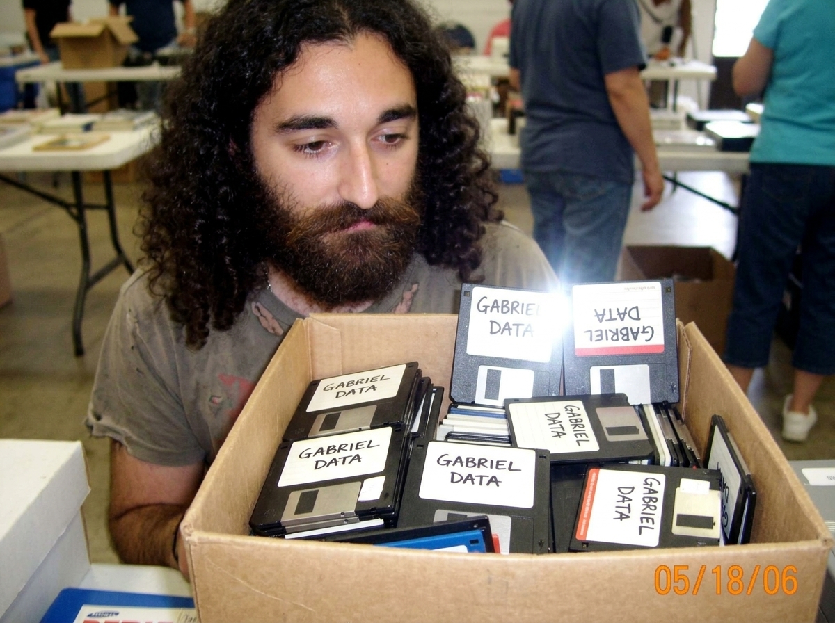 A blurry photo of a cardboard box full of old 3.5-inch floppy disks, all with handwritten labels saying 'GABRIEL DATA' in black sharpie, sitting on a folding table at a yard sale. The photo is taken on a flip phone camera, low resolution, lots of noise.